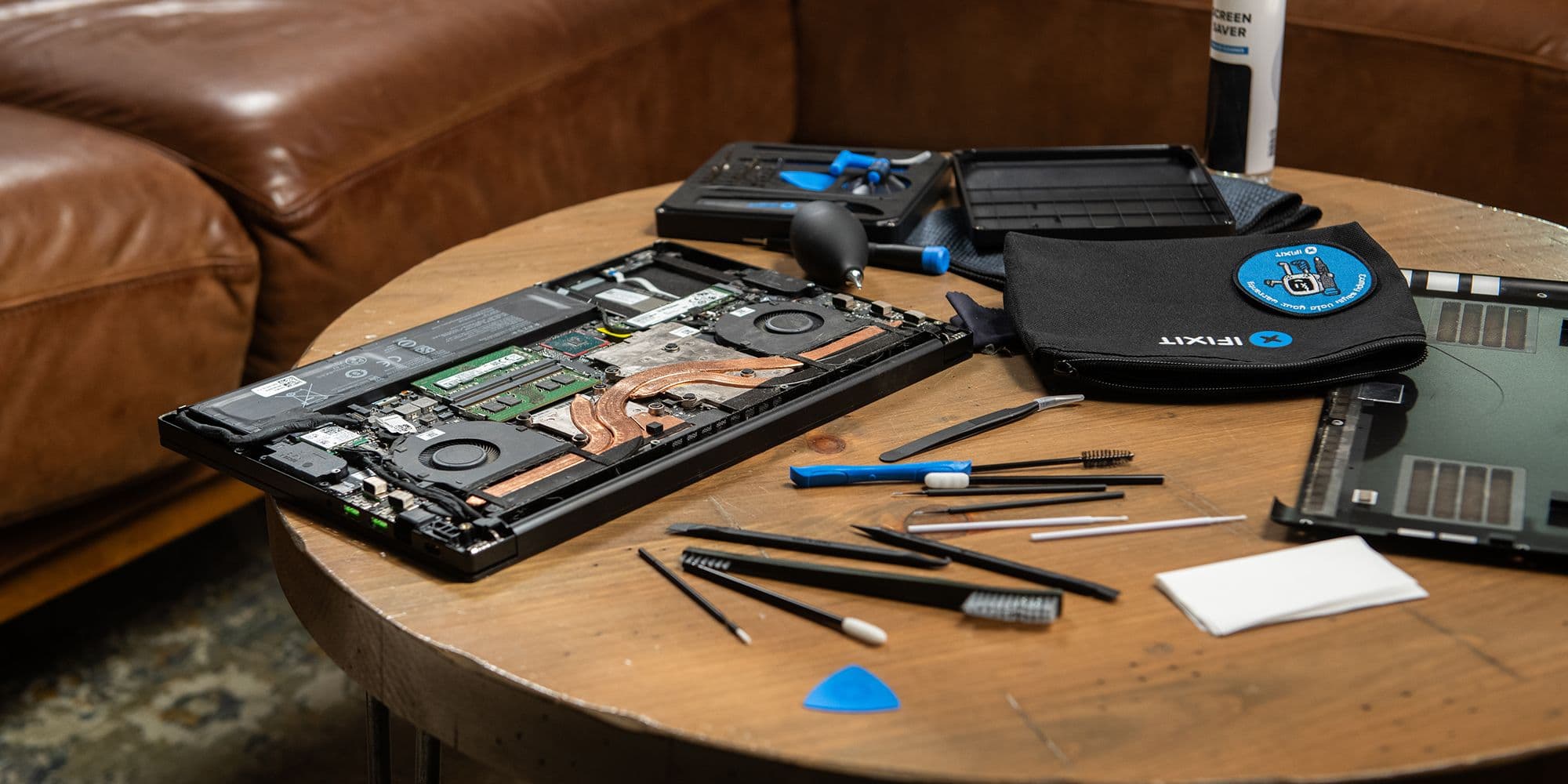 Angled top-down view of an opened laptop on a round wooden table, surrounded by precision tools, brushes, and cleaning accessories. The internal components are exposed while tools and a toolkit are spread out, illustrating an active electronics cleaning and repair setup.